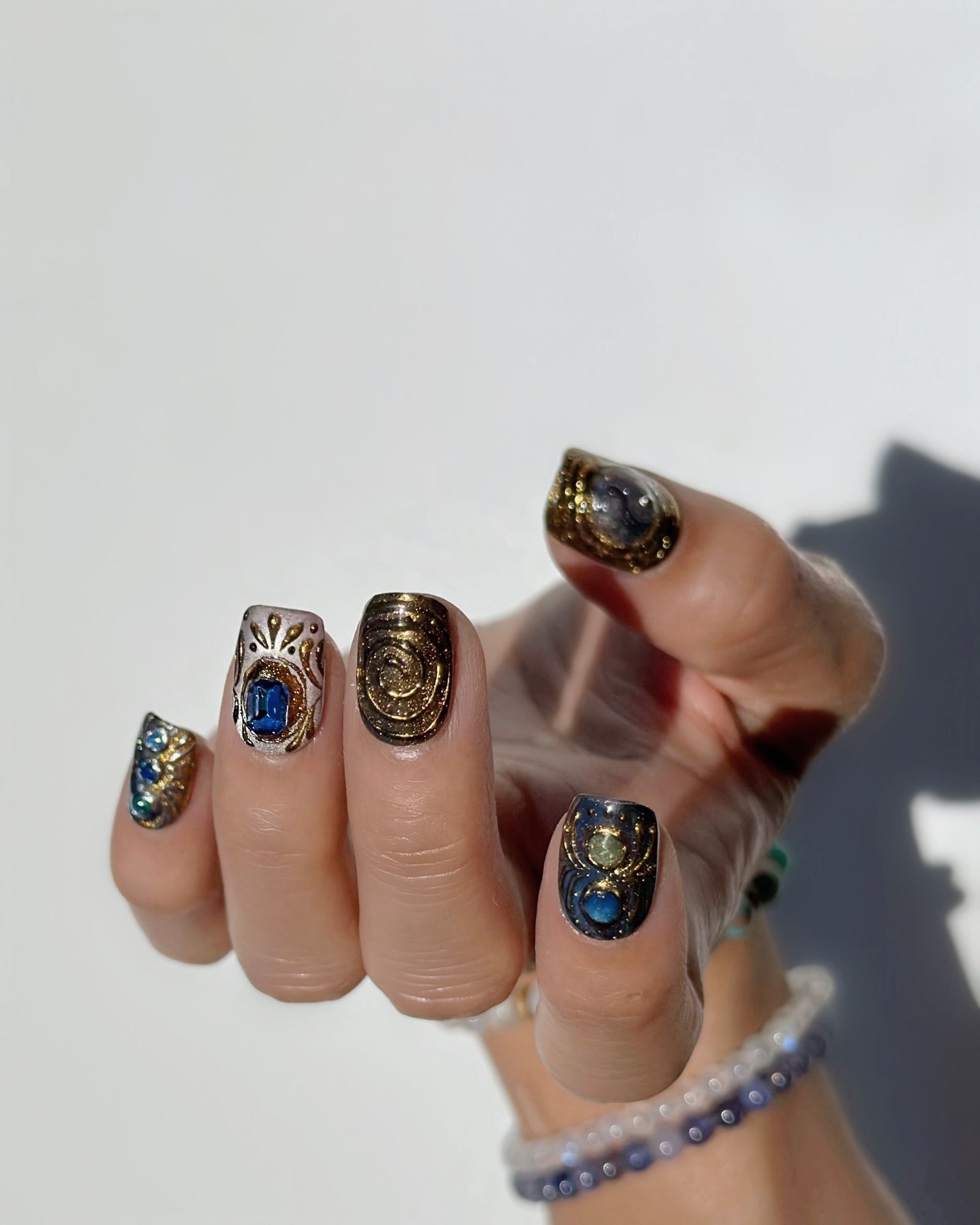 A hand with a palm faced up and fingers curling up to show blue and gold metallic nails with gem and metalswirled details.